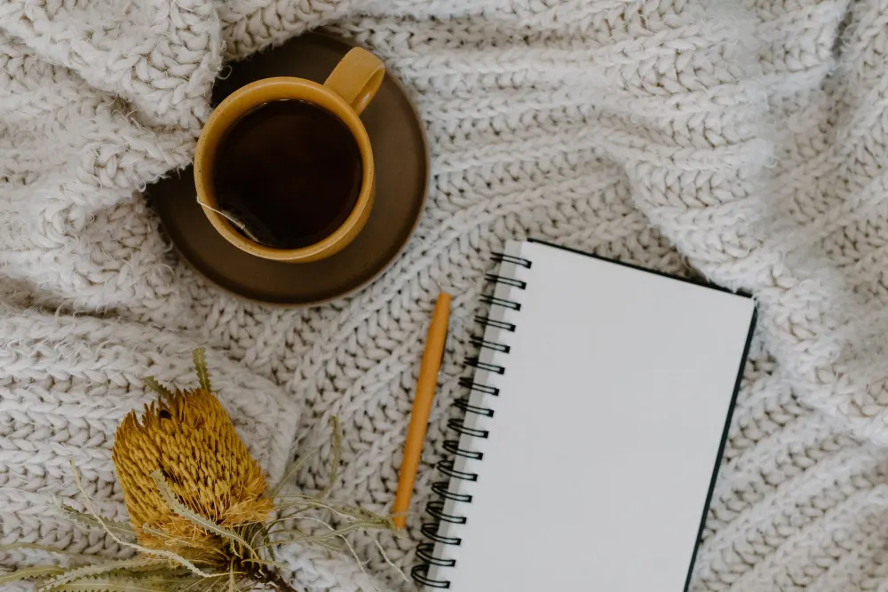 A cup of tea beside an open notebook and pencil on a soft knitted blanket