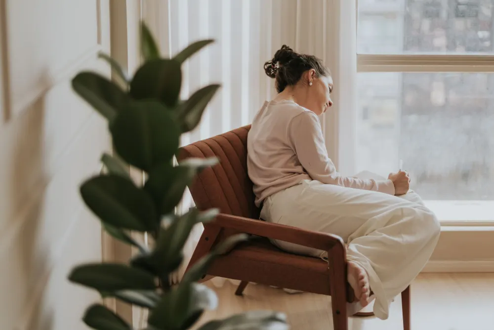A woman sits in a sunlit armchair by the window, writing in a notebook